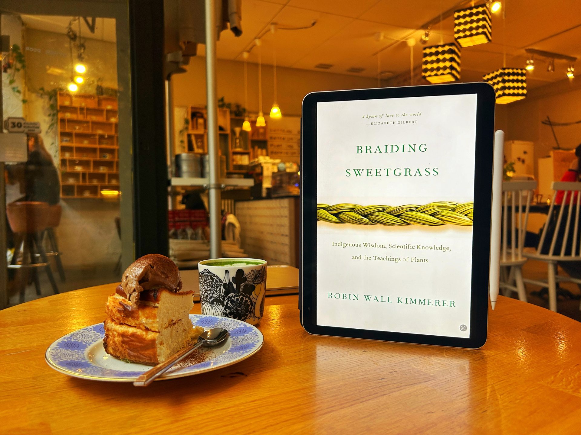 Braiding Sweetgrass Book Cover on the table with a cup of matcha latte and a cake inside a cafe.