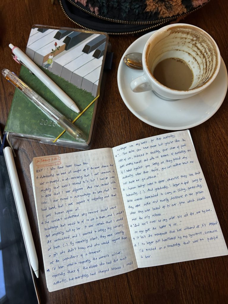 A notebook page with handwritten notes on a wooden table, next to a nearly empty cup of coffee and a planner with pens on top.