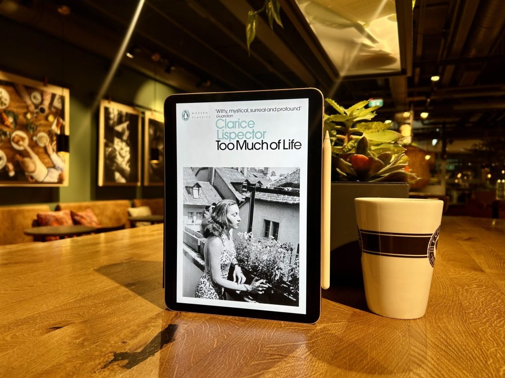 Too Much of Life eBook besides a cup of coffee on a wooden table inside a dimmed light cafe