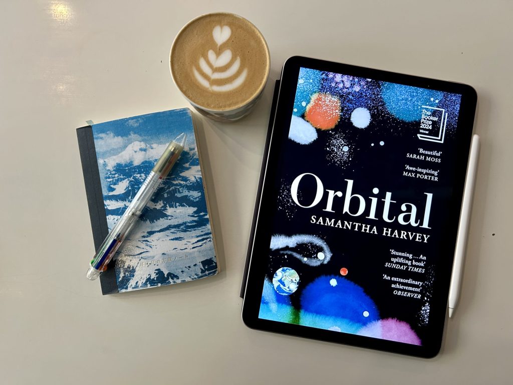 Orbital ebook beside a cup of coffee and a book with a pen on it. All of them are on a white table