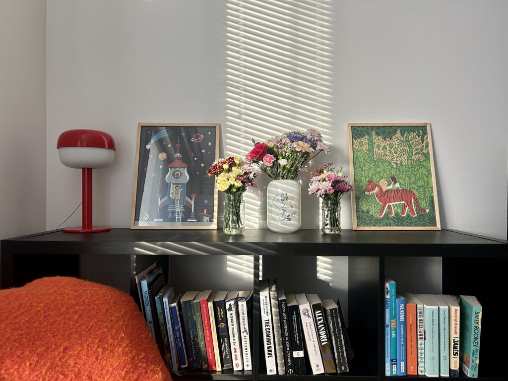 Black bookshelves filled with books and flower vases, decorated with two framed illustrations and a table lamp.