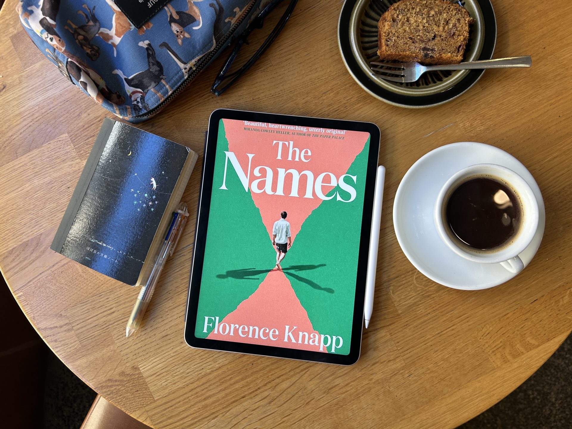 The Names eBook with a cup of black coffee, a slice of banana bread, and a notebook on a wooden table