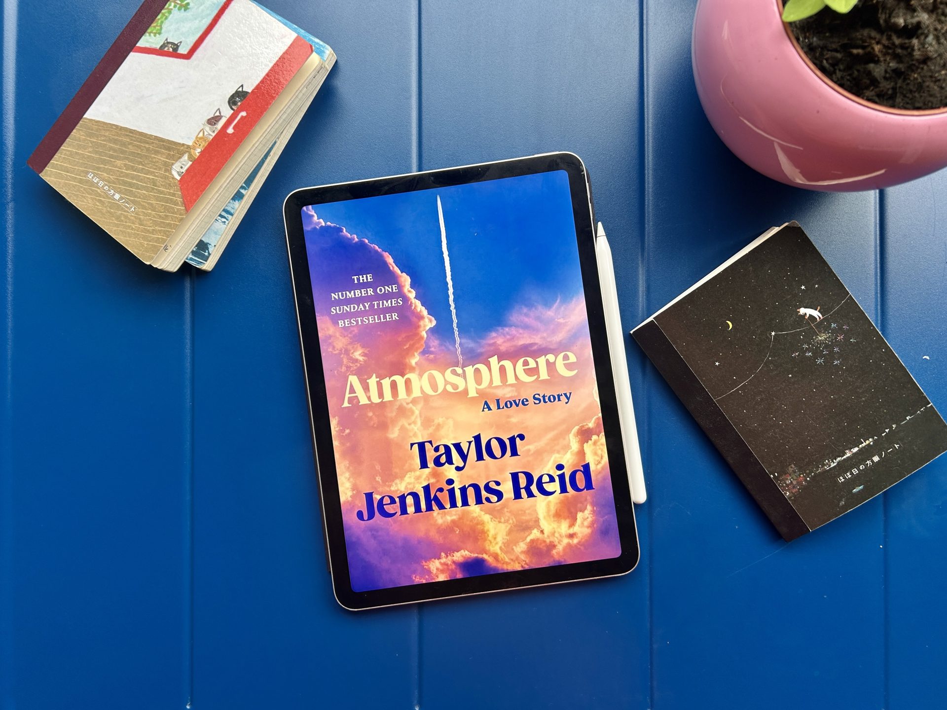 Atmosphere eBook on a solid blue table with some notebooks and a glimpse of plant