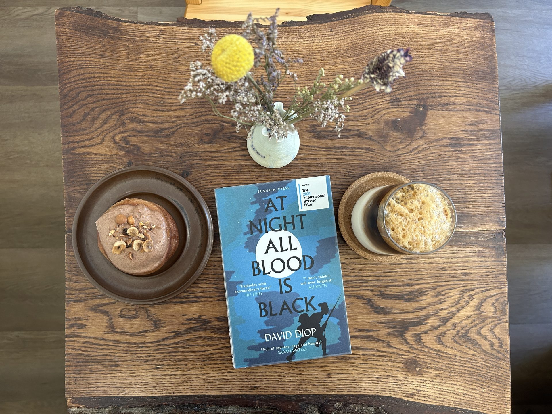 At Night All Blood is Black Book on a wooden table with a glass of iced coffee and a bun